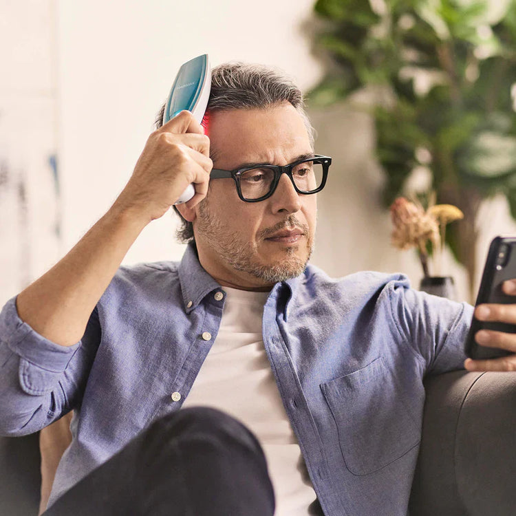 Man using a hairbrush and holding a phone, sitting on a couch indoors.