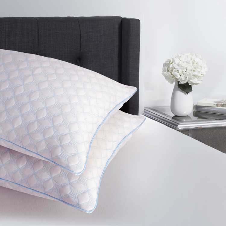 White textured pillow on a bed with a gray headboard and a nightstand with flowers in the background.