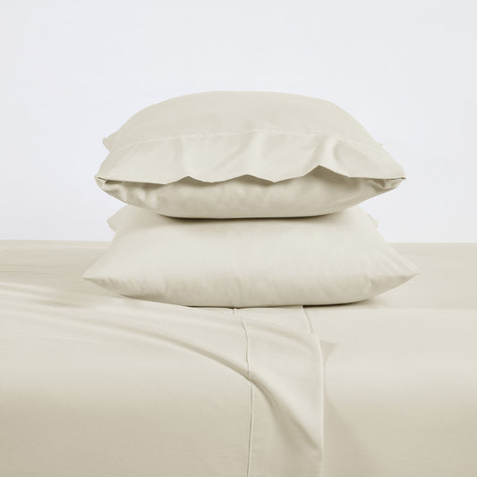 A cream-colored bamboo sheet set on a bed, including a fitted sheet, flat sheet, and pillowcases, with a wooden headboard and houseplants in the room.