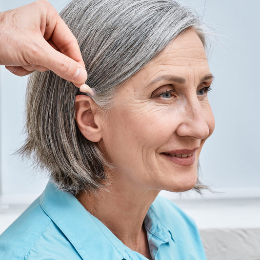 A pair of beige Audien Atom 2 hearing aids with a charging case open and one hearing aid inside, along with a depiction of the other hearing aid outside.