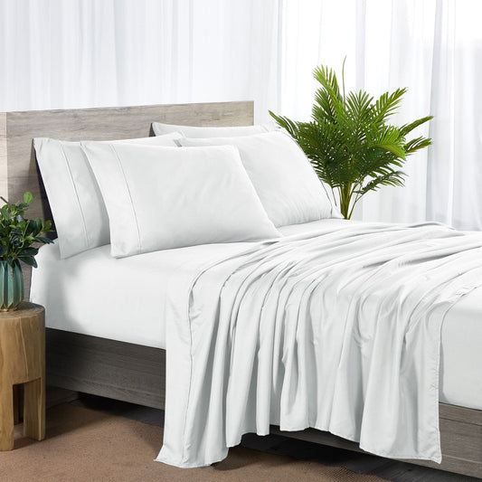 A white 6-piece bamboo sheet set on a bed, including a fitted sheet, flat sheet, and pillowcases, presented in a bright room setting with decorative plants and a nightstand.